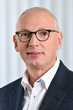 Ein Mann mit kurzen grauen Haaren, Brille und einer Anzugsjacke über einem weißen Hemd, der in die Kamera schaut und leicht lächelt.